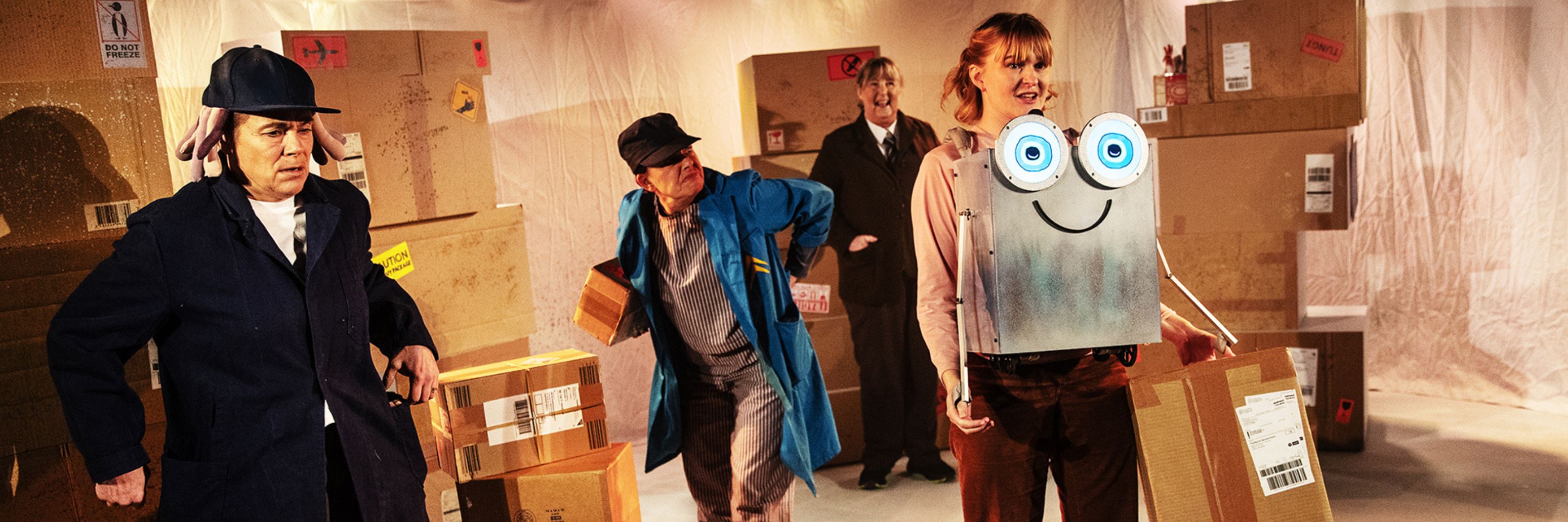 En skådespelare har en robot på magen och bär lådor. Till vänster syns två lagerarbetare i keps som ser missnöjda ut. I bakgrunden, bland lådor, står en chef i kostym och ler.